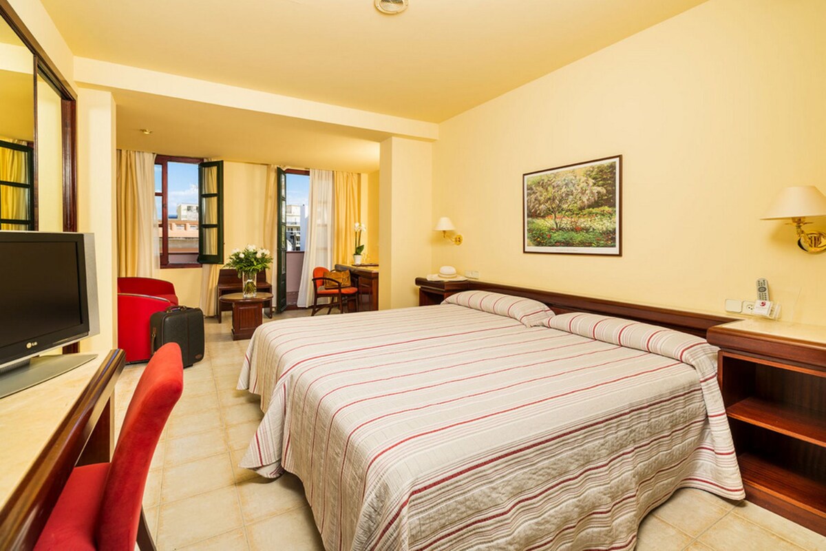 A hotel room with a large bed, a television, a desk with a chair, a red chair, a lamp, and a painting on the wall.