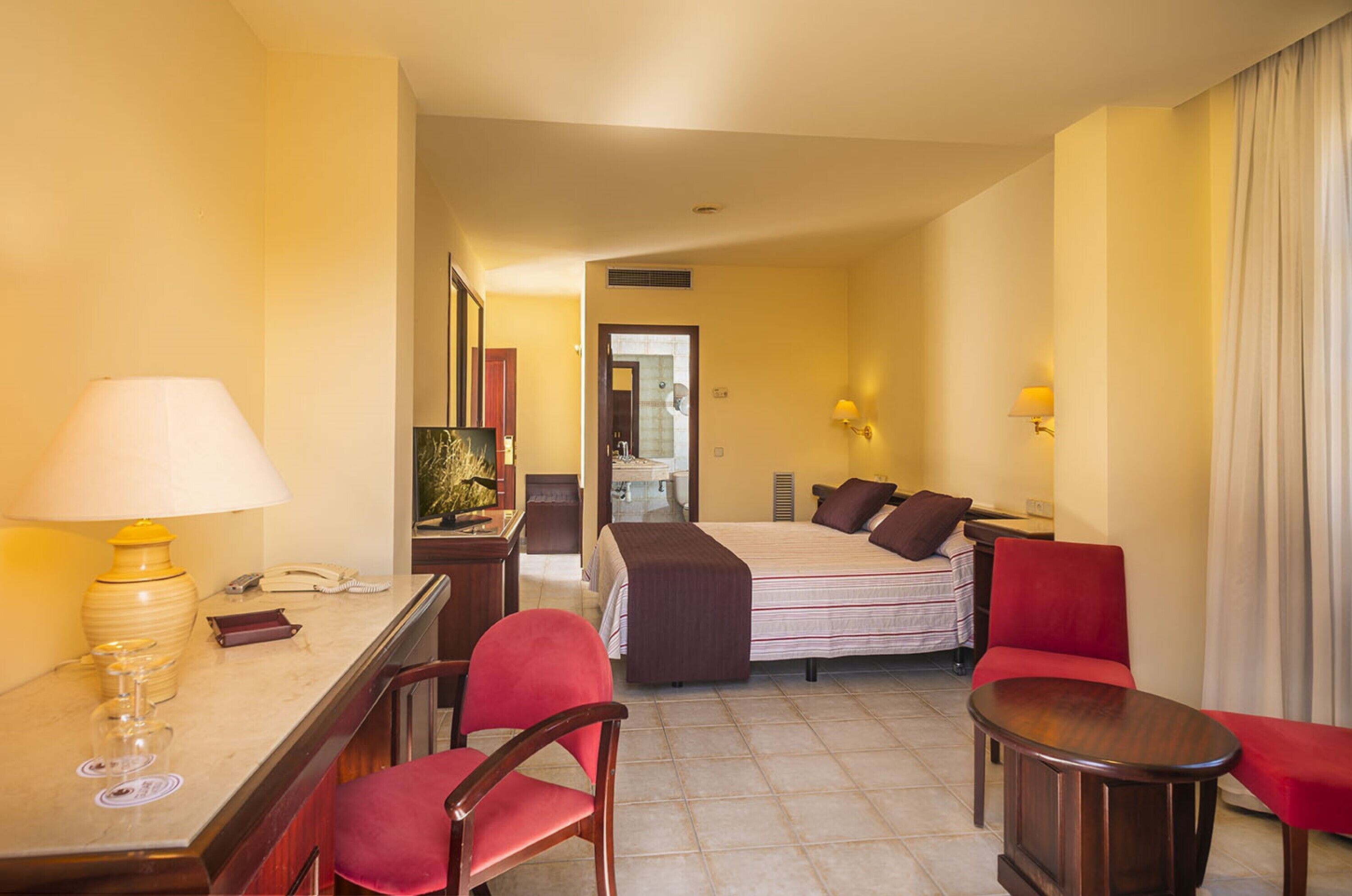 Una habitación de hotel con cama, escritorio con lámpara, una silla, una mesita y un espejo.