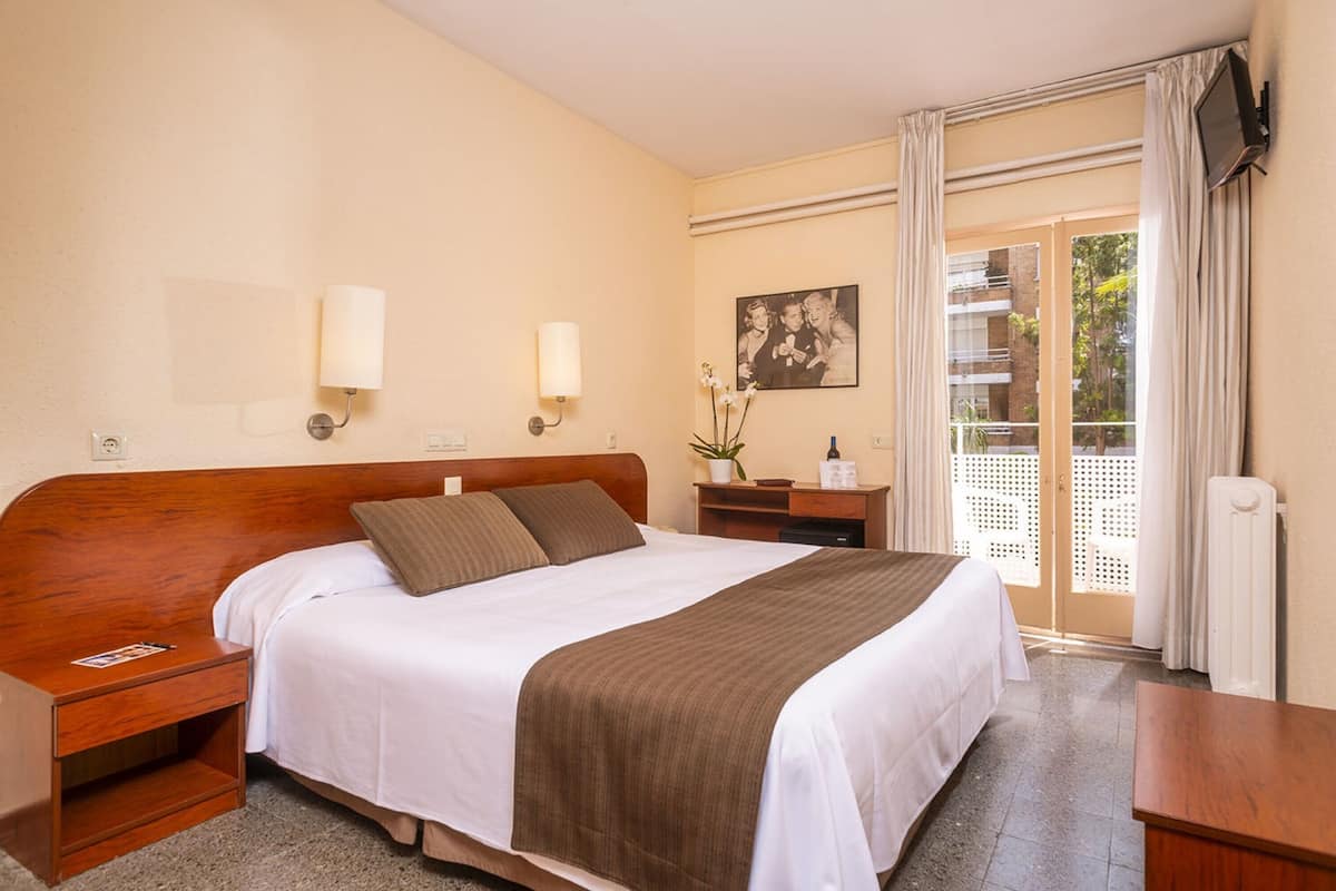 A hotel room with a large bed, a nightstand, a bedside table, a potted plant, a painting on the wall, and a sliding glass door leading to a balcony.