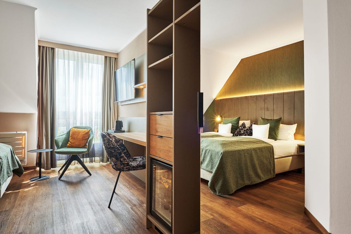 Een moderne hotelkamer met een houten vloer, een groene fauteuil, een bureau met stoel, een bed met een groene deken en een groot raam met gordijnen.