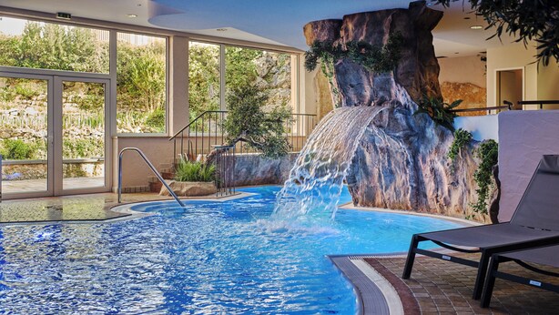 Piscina interior con caída de agua, rodeada de amplios ventanales y suelo de ladrillo.