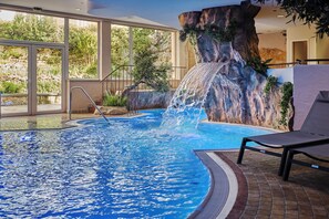 Une piscine intérieure agrémentée d’une cascade, entourée de grandes fenêtres et dotée d’un sol en briques.