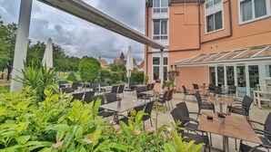 Un café al aire libre con mesas y sillas, rodeado de follaje y un edificio con techo de cristal.