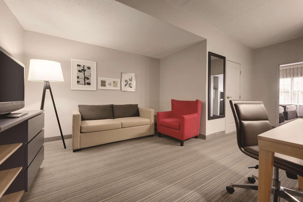 A hotel room with a sofa, a red armchair, a desk with a chair, a lamp, and framed pictures on the wall.