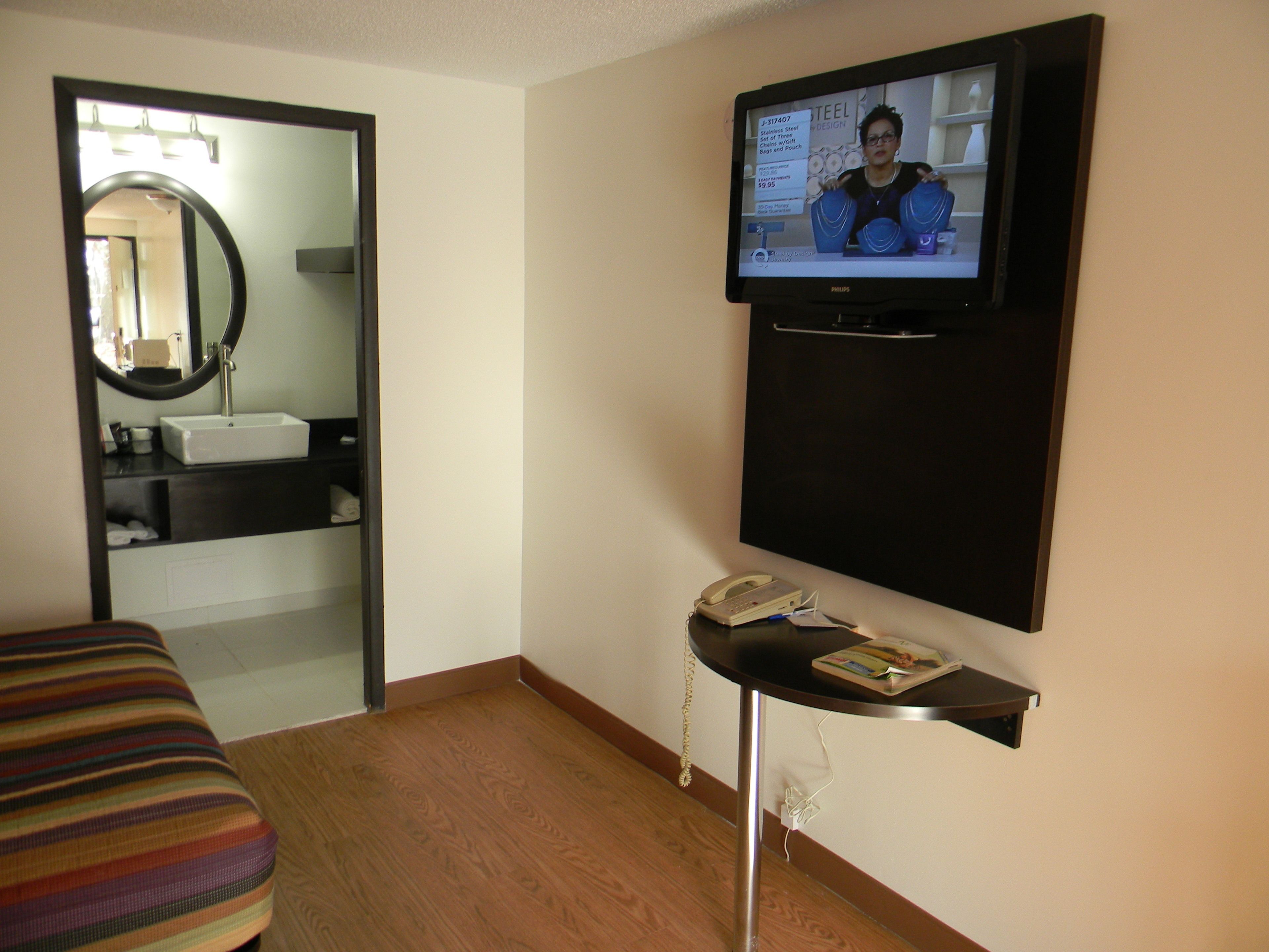 Habitación de hotel con televisión montada en la pared, mesita de noche con teléfono y revista, cama a rayas, baño con lavamanos y espejo, y suelo de madera.