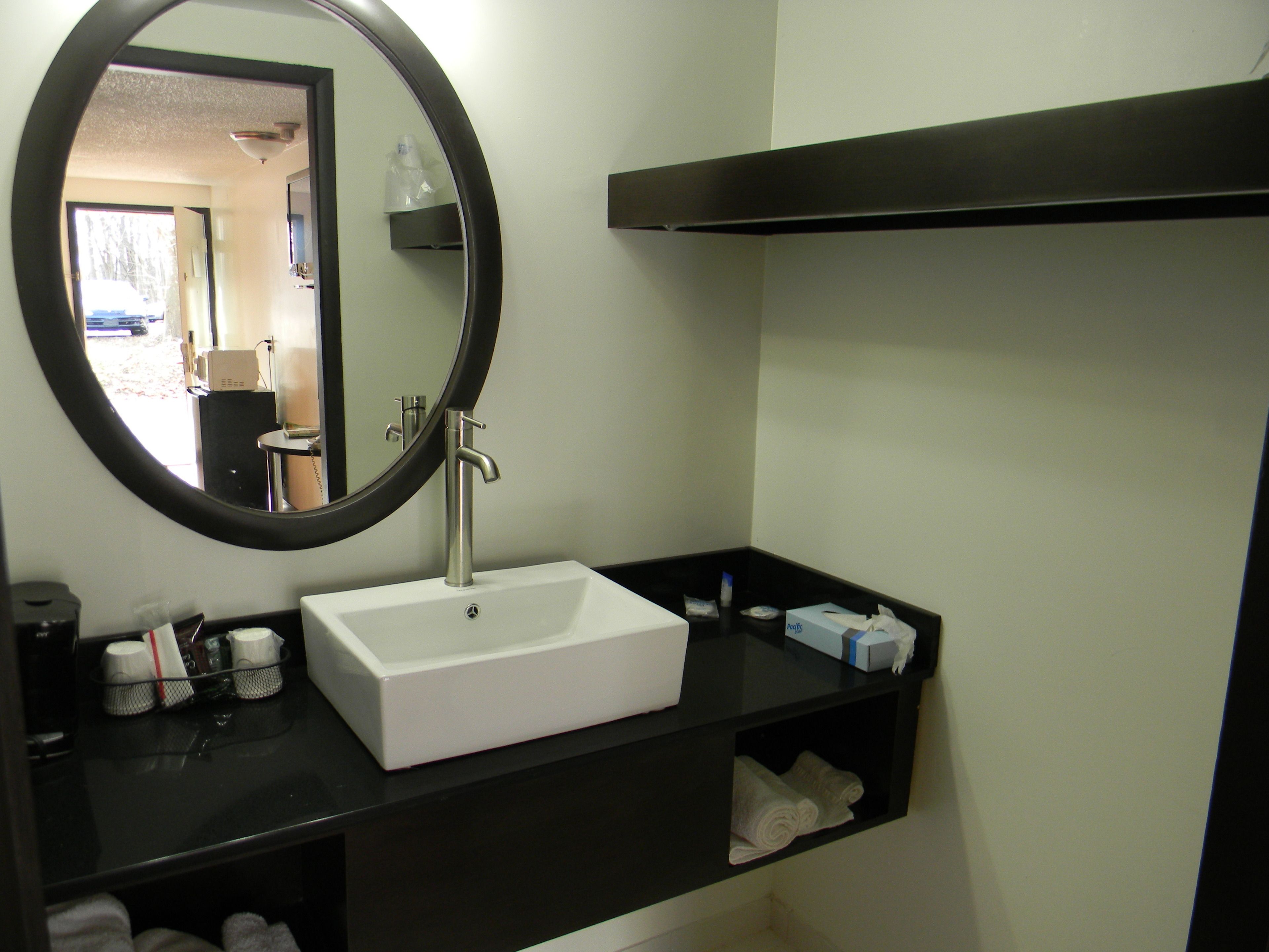 A bathroom with a white sink, black countertop, and a round mirror. There is a towel rack with towels and a soap dispenser on the countertop.