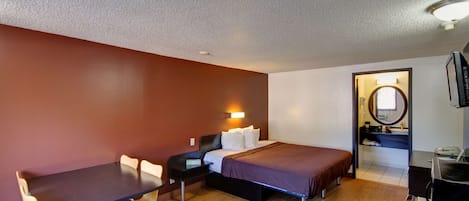 Habitación de hotel pequeña con cama, escritorio y silla. Hay un espejo y una mesita de noche con lámpara.