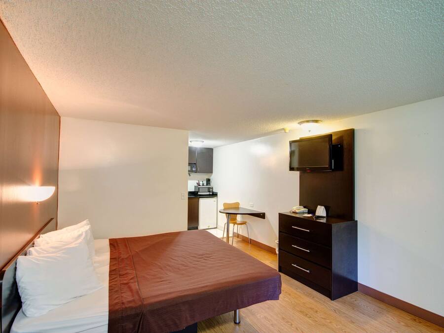 Habitación de hotel pequeña con cama, escritorio, silla, kitchenette y televisor montado en la pared.