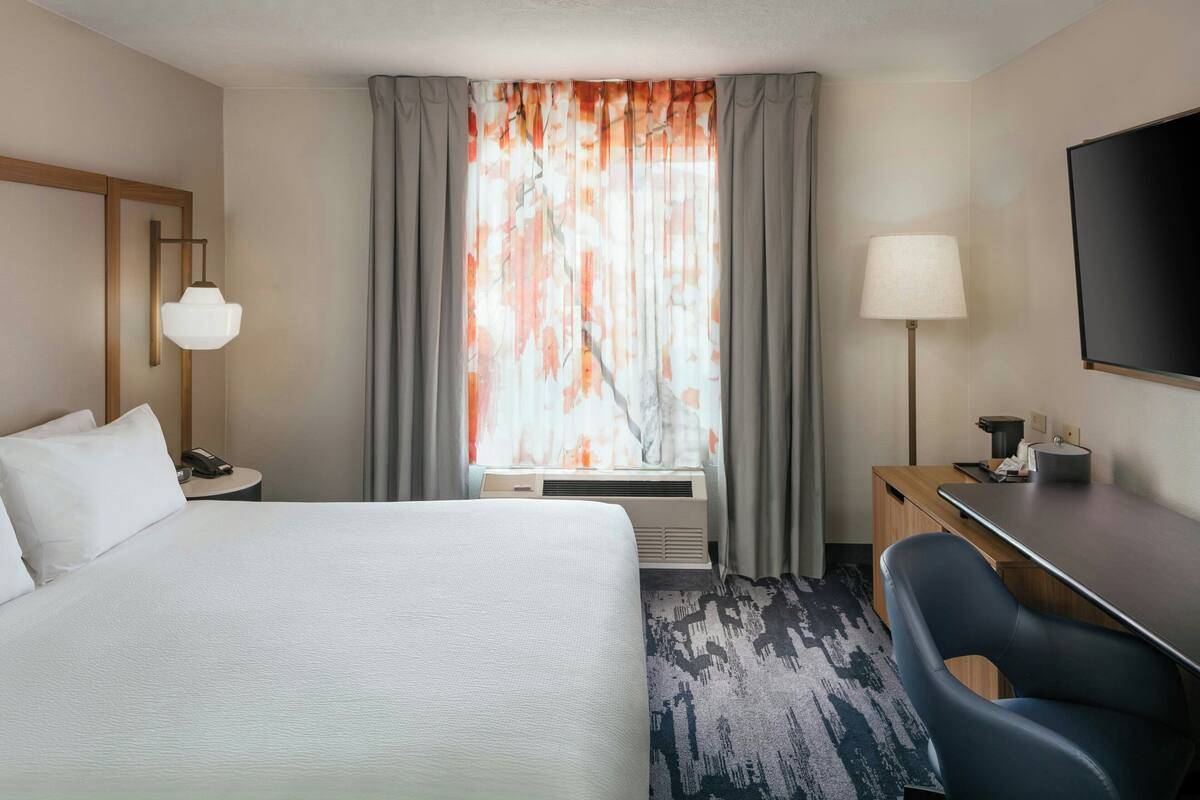 Une chambre d’hôtel équipée d’un lit, d’un bureau, d’une chaise, d’une lampe et d’une grande fenêtre avec des rideaux.