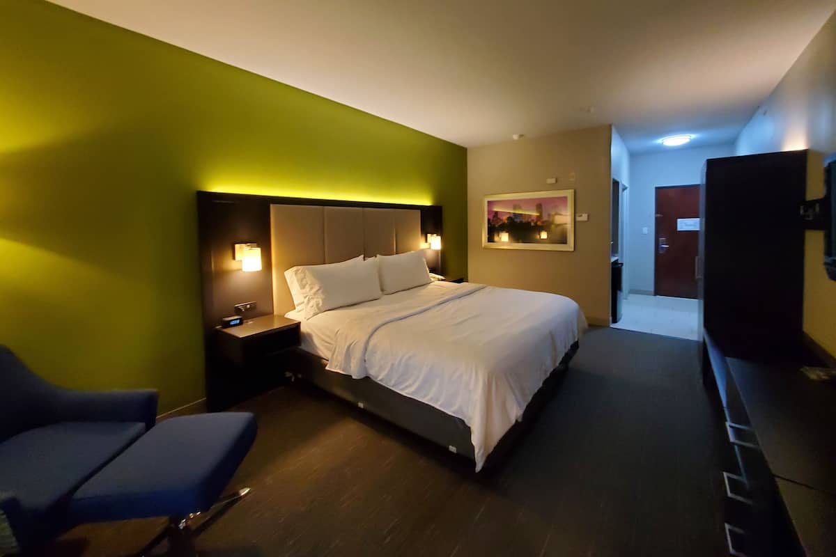 A hotel room with a bed, bedside table, lamp, chair, and a painting on the wall.