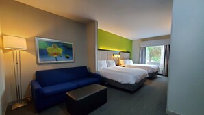 Ett hotellrum med en blå soffa, två sängar, ett litet bord, en tavla med blommor och ett fönster med gardiner.