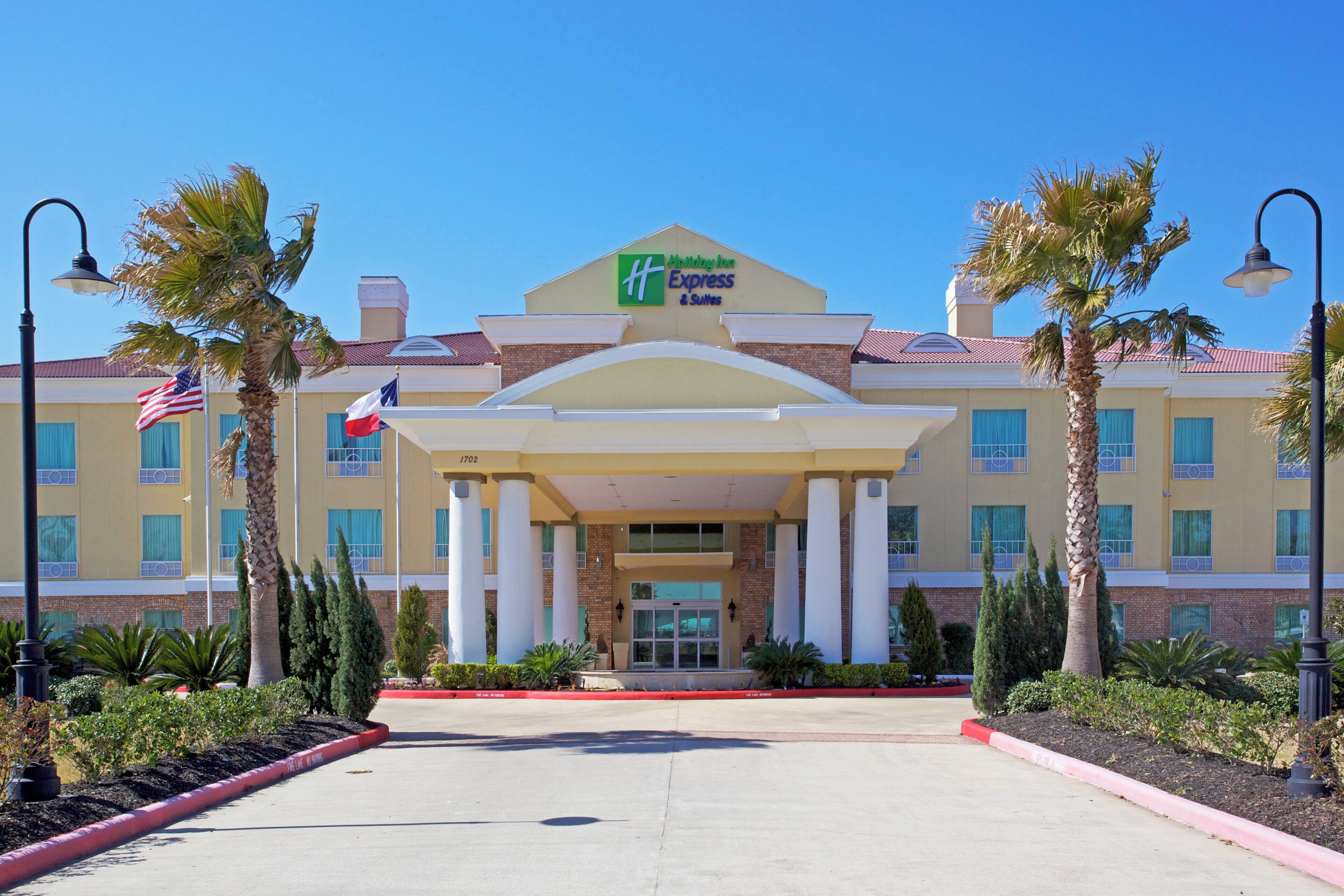 A Holiday Inn Express & Suites hotel with a central entrance, palm trees, and a clear blue sky.