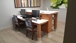 A workspace with two desks, each with a computer and a printer. There are two chairs, one with a patterned backrest. The room has a light-colored floor and walls with artwork on the upper wall.
