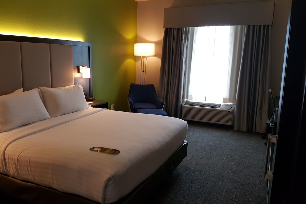 A hotel room with a large bed, a nightstand, a chair, a lamp, and a window with curtains.