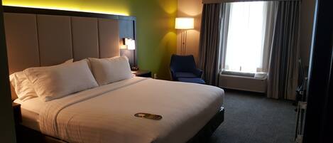 Una habitación de hotel con una cama grande, una mesita de noche, una silla, una lámpara y una ventana con cortinas.