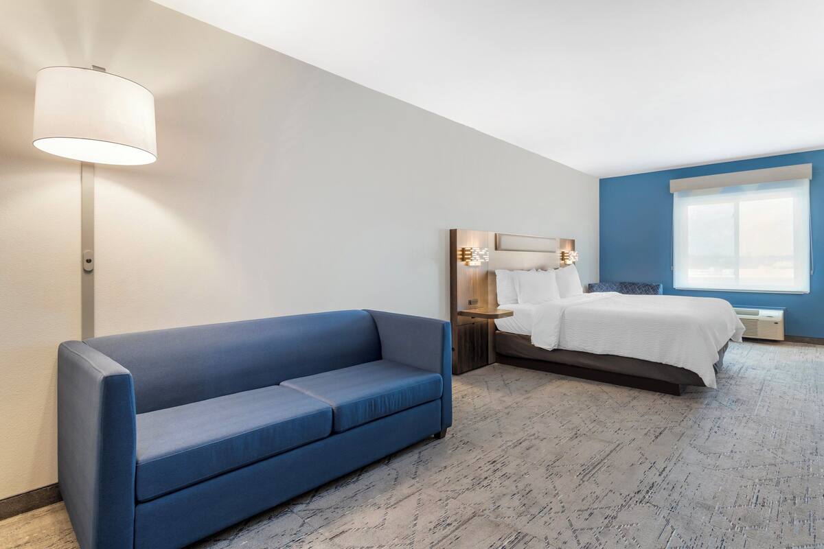 Une chambre d’hôtel moderne avec un canapé bleu, un lit avec une literie blanche, une table de chevet et une grande fenêtre.