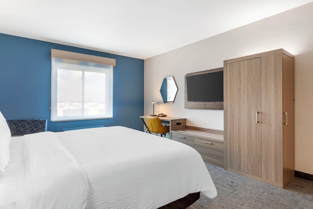 Une chambre d’hôtel comprenant un lit, un bureau avec une chaise, une armoire, un téléviseur fixé au mur et une fenêtre avec des stores.