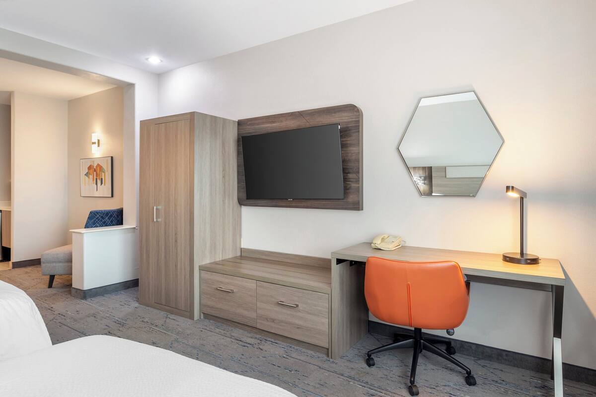 Une chambre d’hôtel comprenant un lit, un bureau avec une chaise orange, une armoire, un téléviseur à écran plat et un miroir fixé au mur.