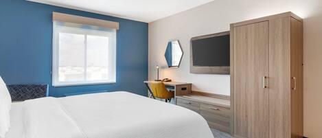Habitación de hotel con cama, escritorio con silla, armario, televisor montado en la pared y ventana con persianas.