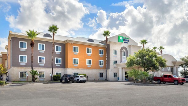 A multi-story hotel with a brown and beige exterior, palm trees, and a parking lot with several vehicles. - Holiday Inn Express & Suites Alice by IHG (Alice)