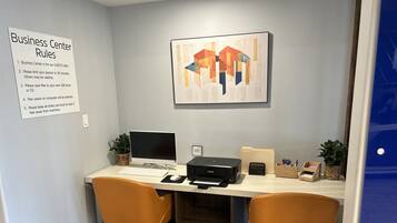 A business center desk with a computer, printer, and office supplies. There is a sign listing business center rules and a framed abstract painting on the wall.