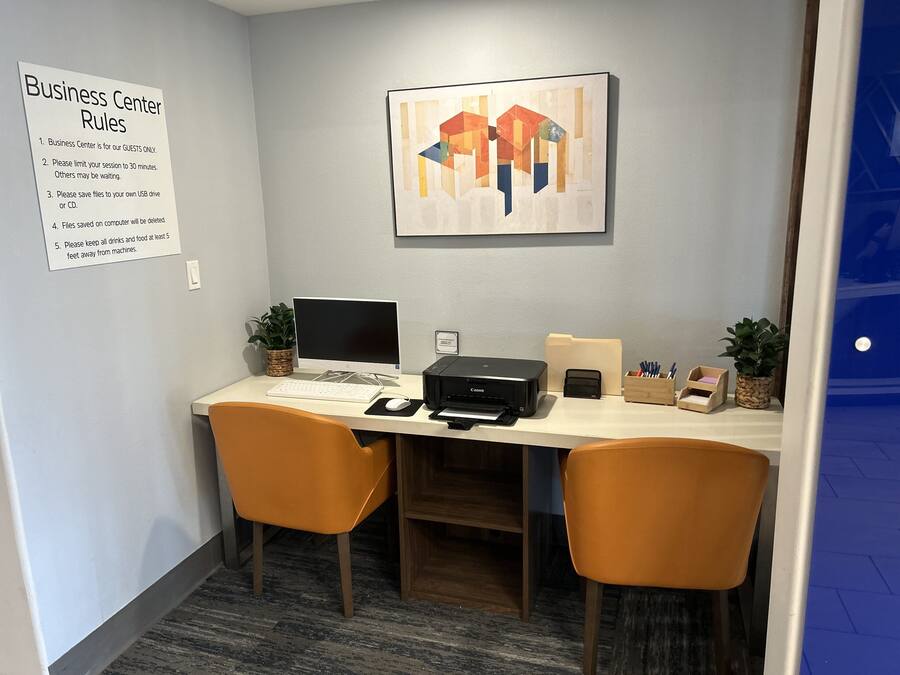 A business center desk with a computer, printer, and office supplies. There is a sign listing business center rules and a framed abstract painting on the wall.