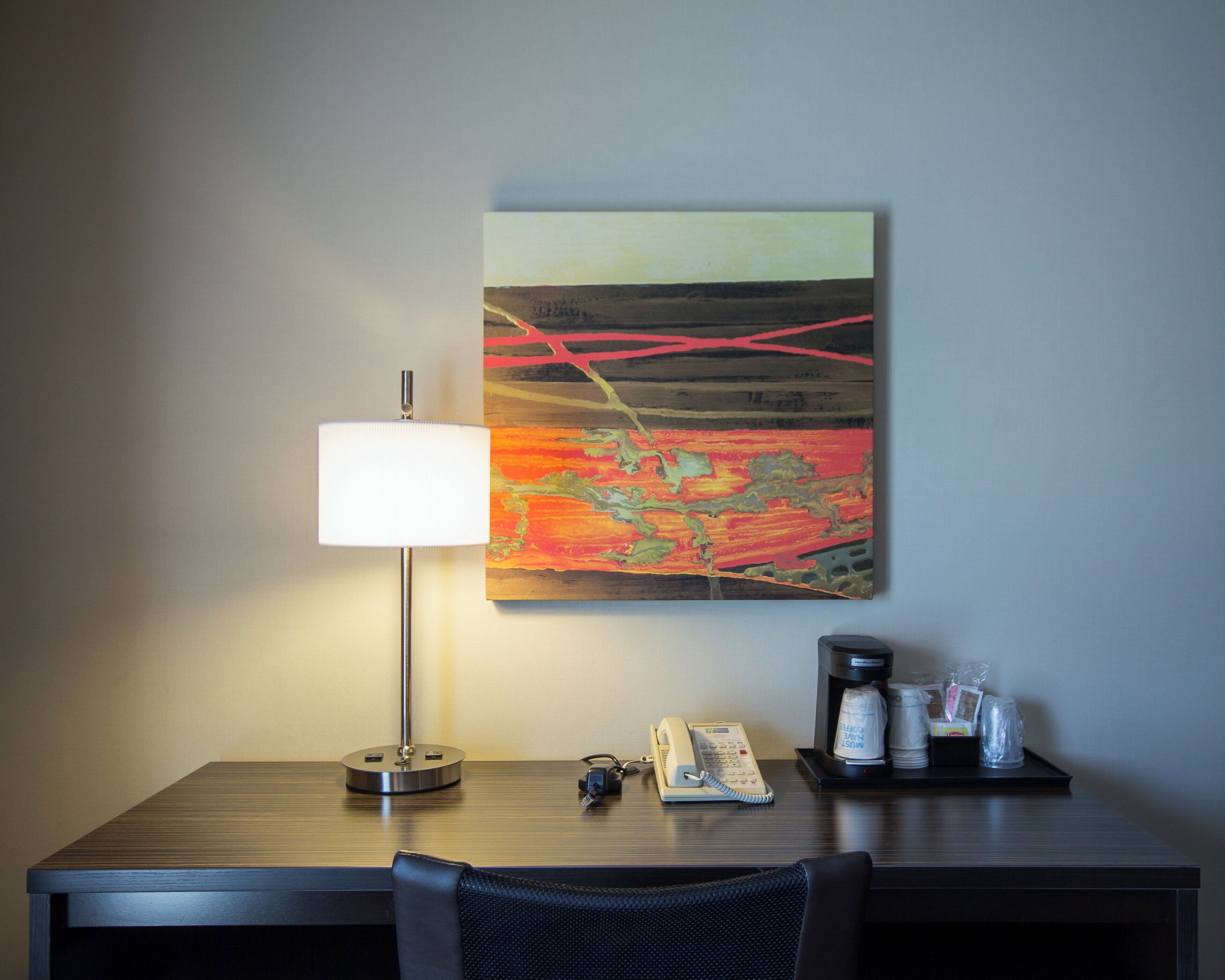 Een hotelkamer met een bureau, lamp, schilderij, telefoon en koffiezetapparaat.