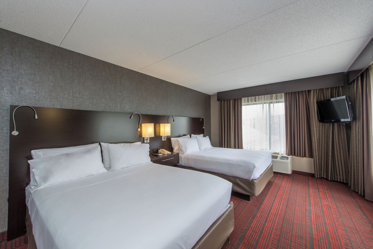 Une chambre d’hôtel avec deux lits, un téléviseur à écran plat, une fenêtre avec des rideaux et un sol recouvert d’une moquette rouge.
