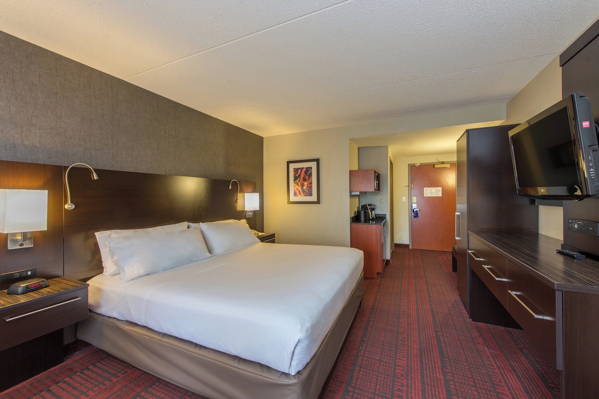 A hotel room with a large bed, a desk with a lamp, a television, and a kitchenette area.