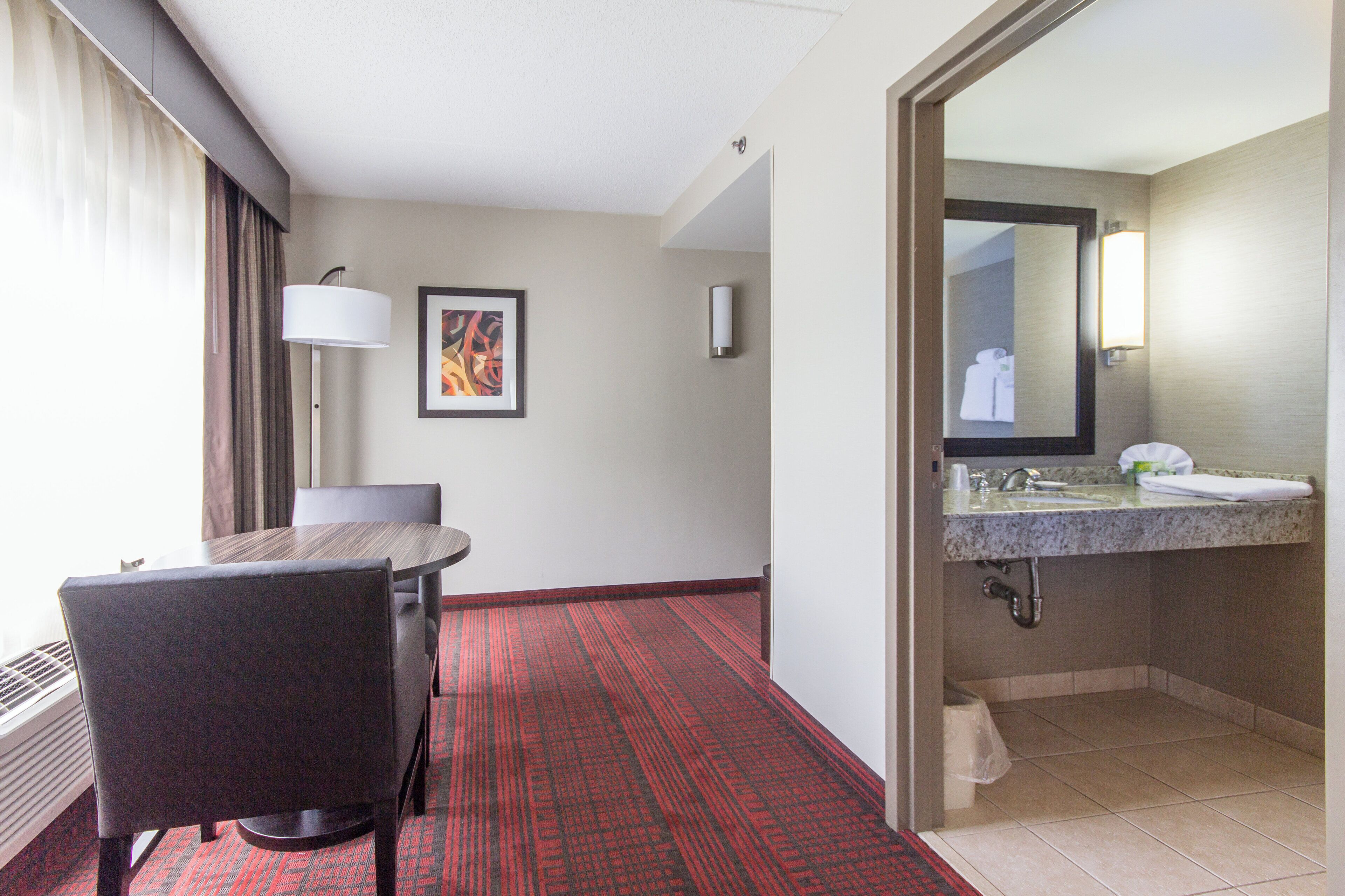 Habitación de hotel con una mesa de comedor, una silla, alfombra roja con diseño, un cuadro abstracto enmarcado, una lámpara de pared y un baño con lavamanos y espejo.