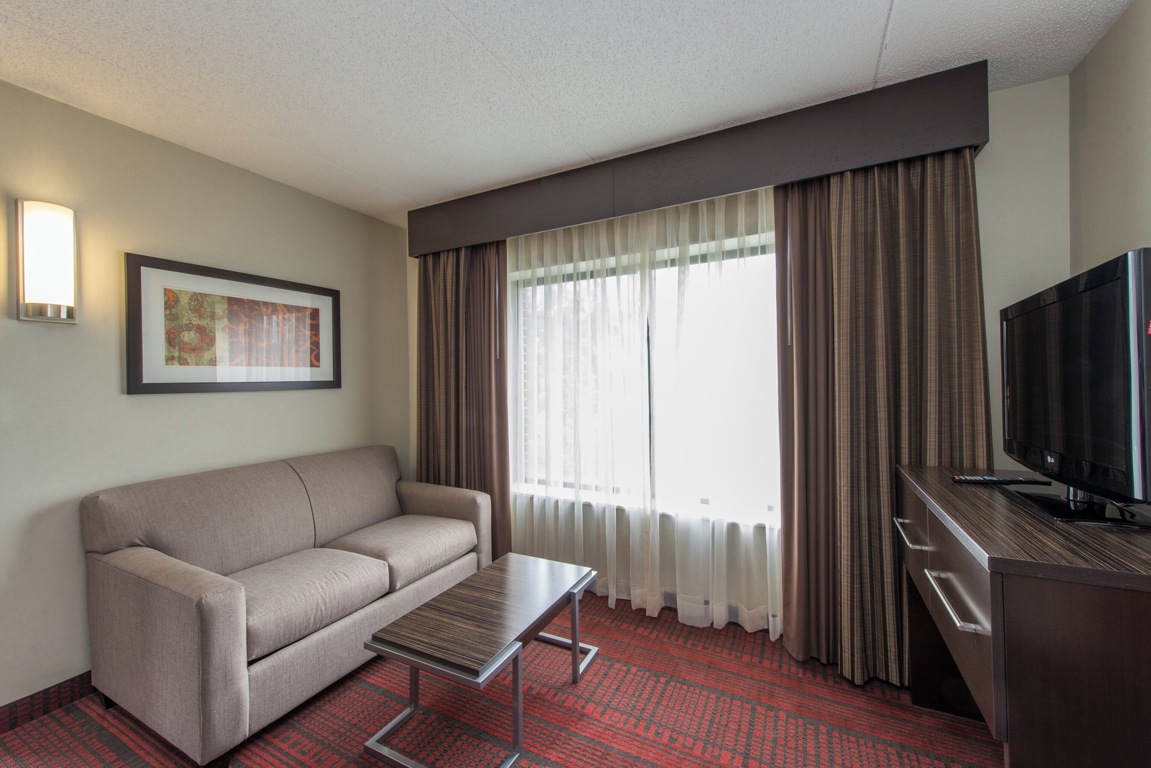 Una habitación de hotel con sofá, una mesita, televisión y un amplio ventanal con cortinas.