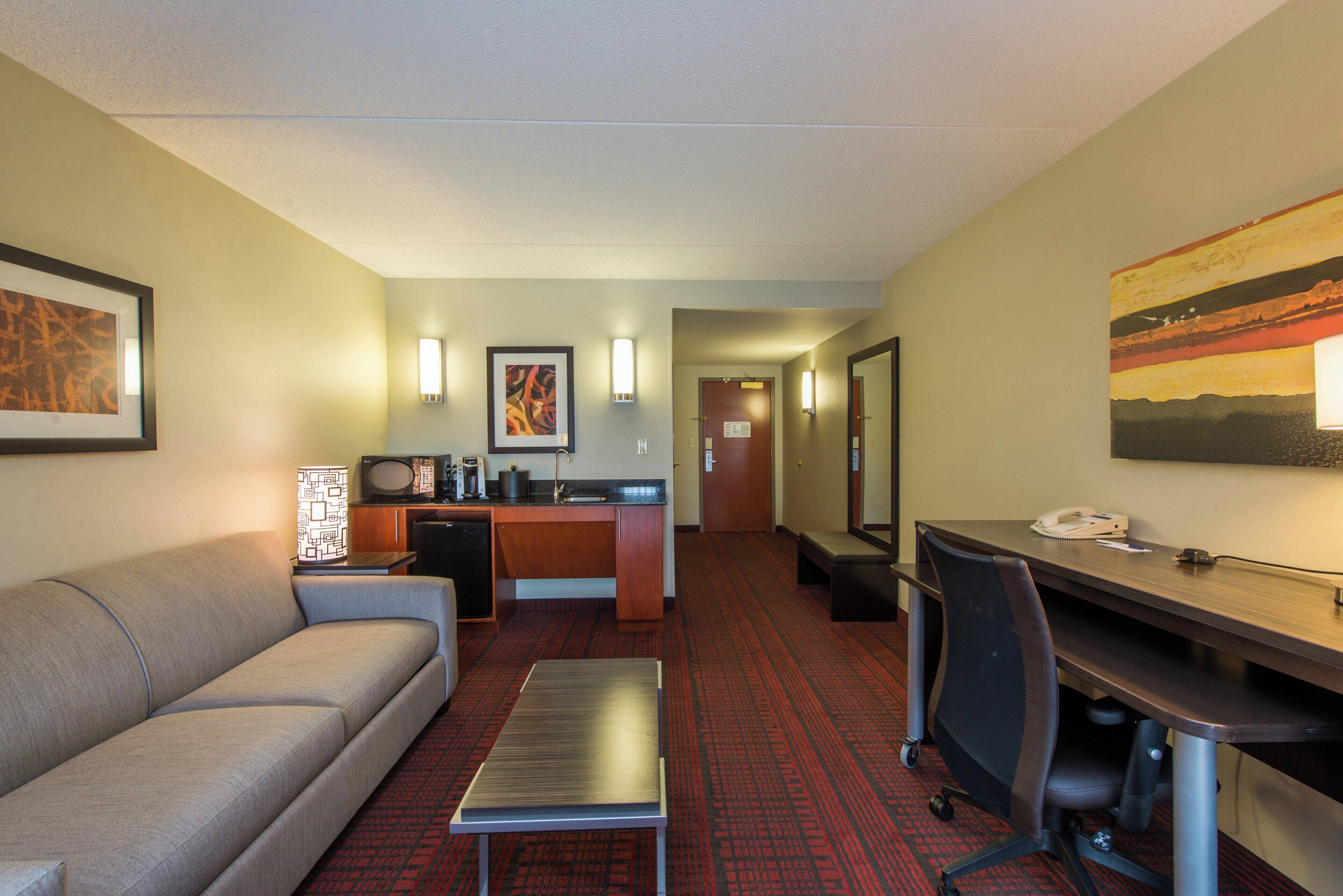 A hotel room with a sofa, a desk, a small kitchenette, and a hallway leading to a door.