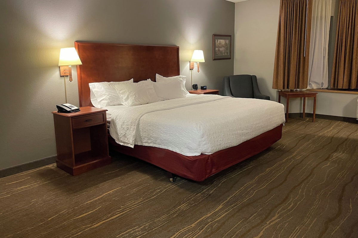 A hotel room with a large bed, two bedside lamps, a nightstand, a chair, a small table, and a window with curtains.