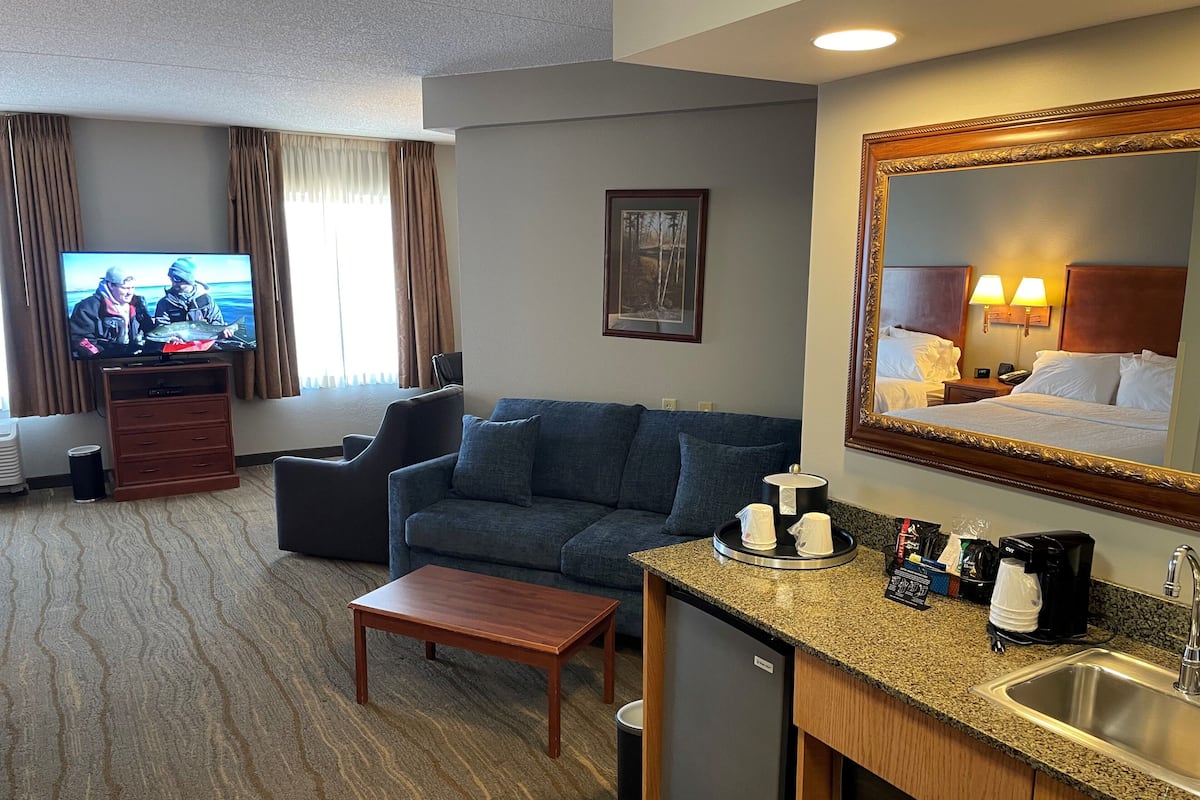 A hotel room with a TV, sofa, coffee table, kitchenette, and a large mirror.