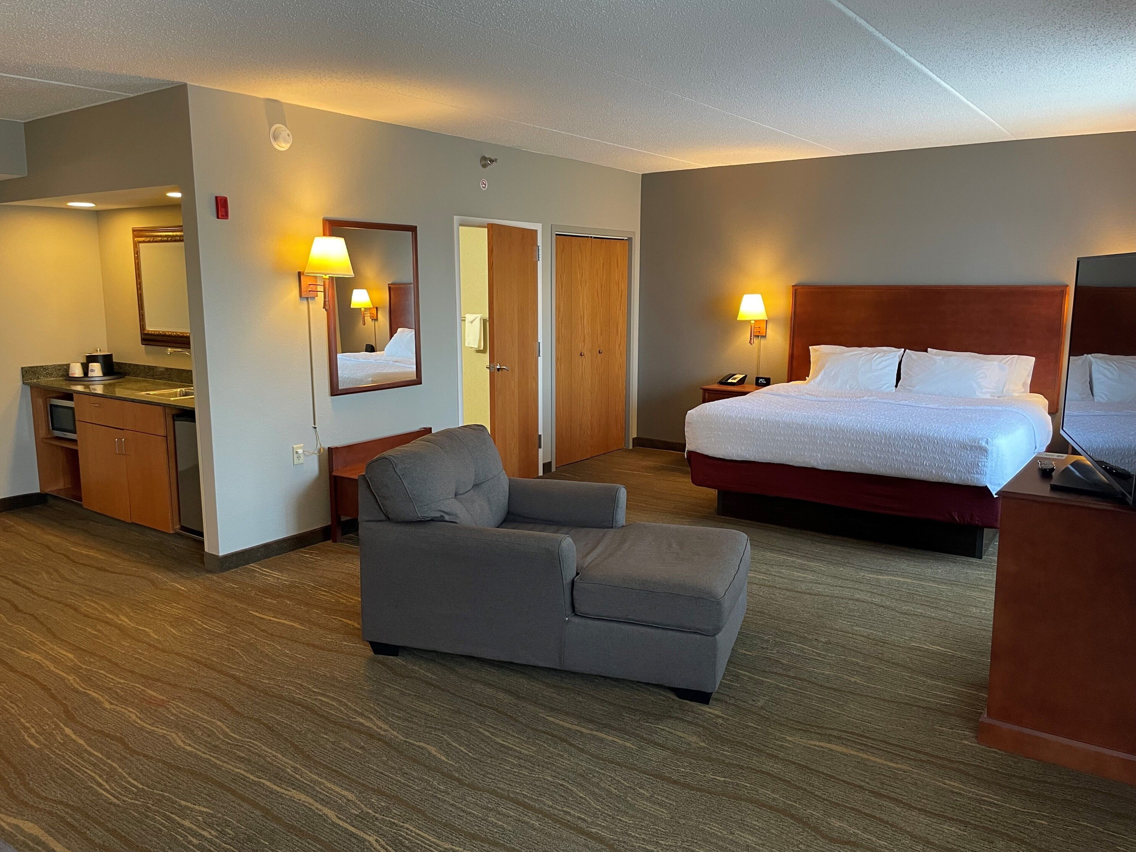 A hotel room with a bed, a chair, a desk with a lamp, a mirror, and a small kitchenette.
