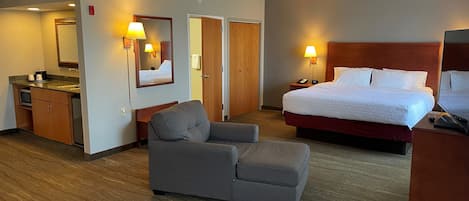 A hotel room with a bed, a chair, a desk with a lamp, a mirror, and a small kitchenette.