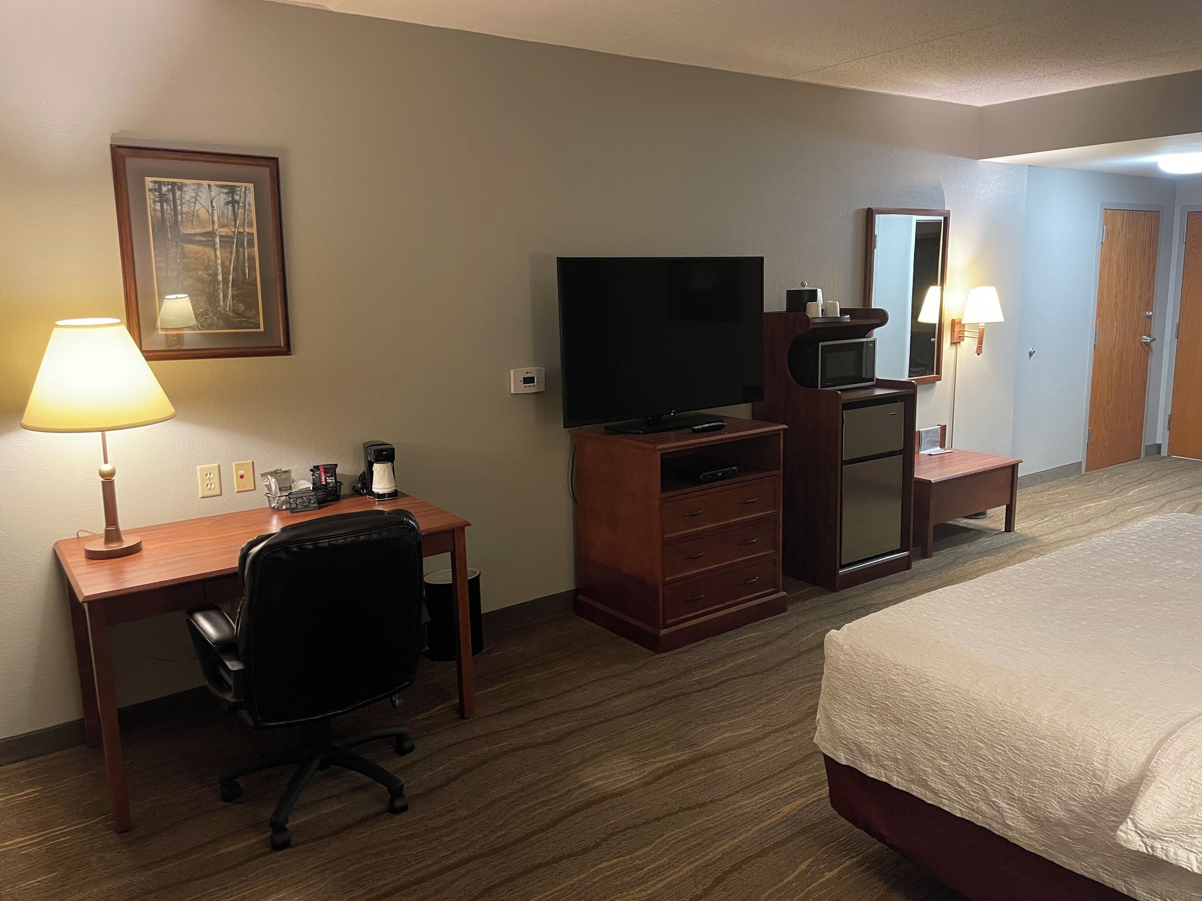 A hotel room with a bed, a desk with a chair, a television, a coffee maker, a mirror, and a lamp.