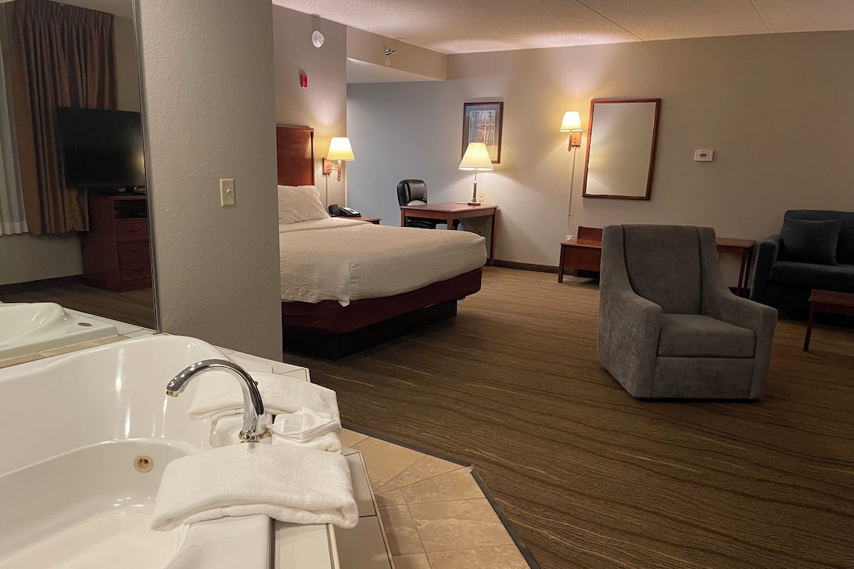 A hotel room with a large bed, a sitting area with a chair, a desk, a lamp, and a television.