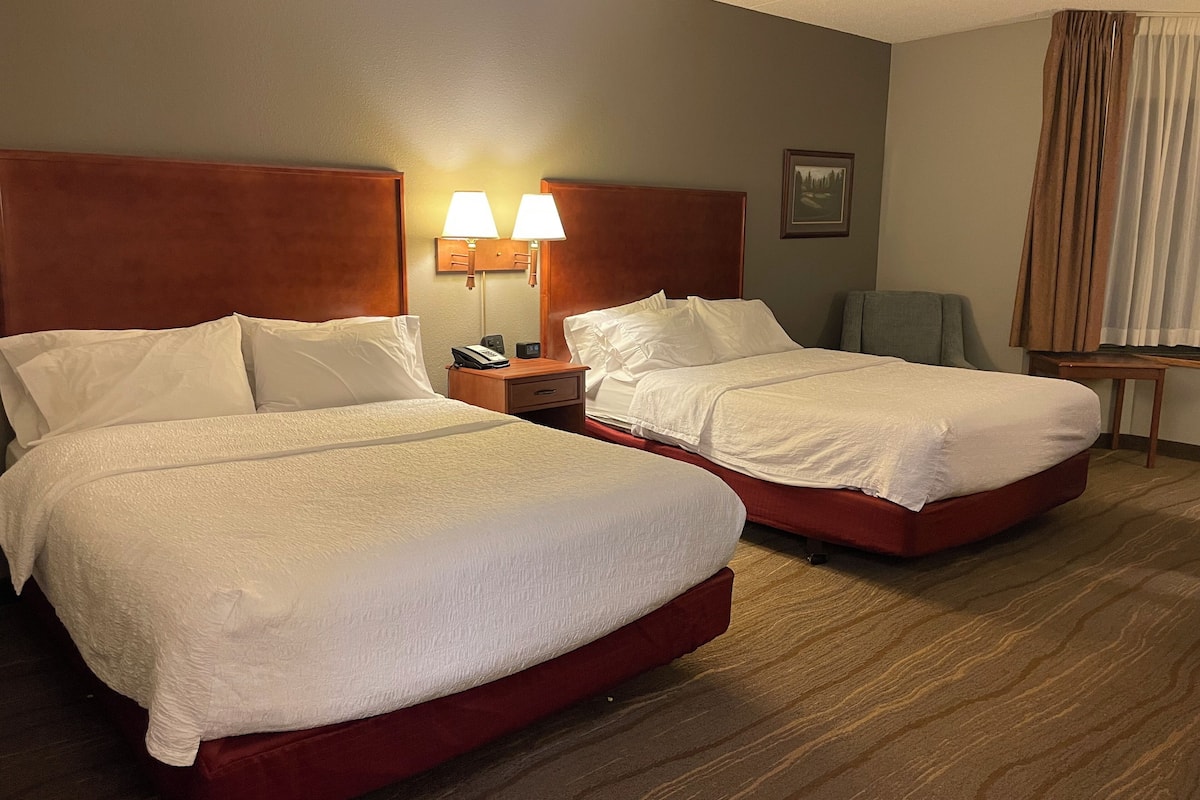 A hotel room with two beds, a nightstand with a lamp, a chair, and a small table.
