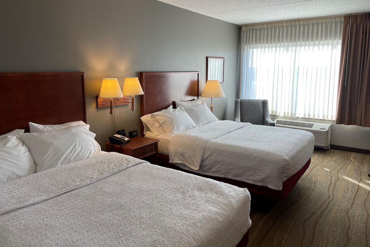 A hotel room with two beds, a nightstand with lamps, a chair, and a large window with curtains.