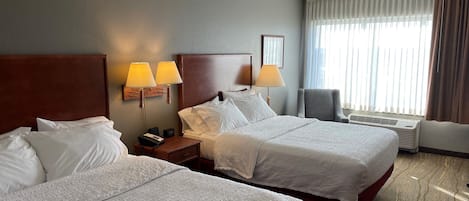 A hotel room with two beds, a nightstand with lamps, a chair, and a large window with curtains.