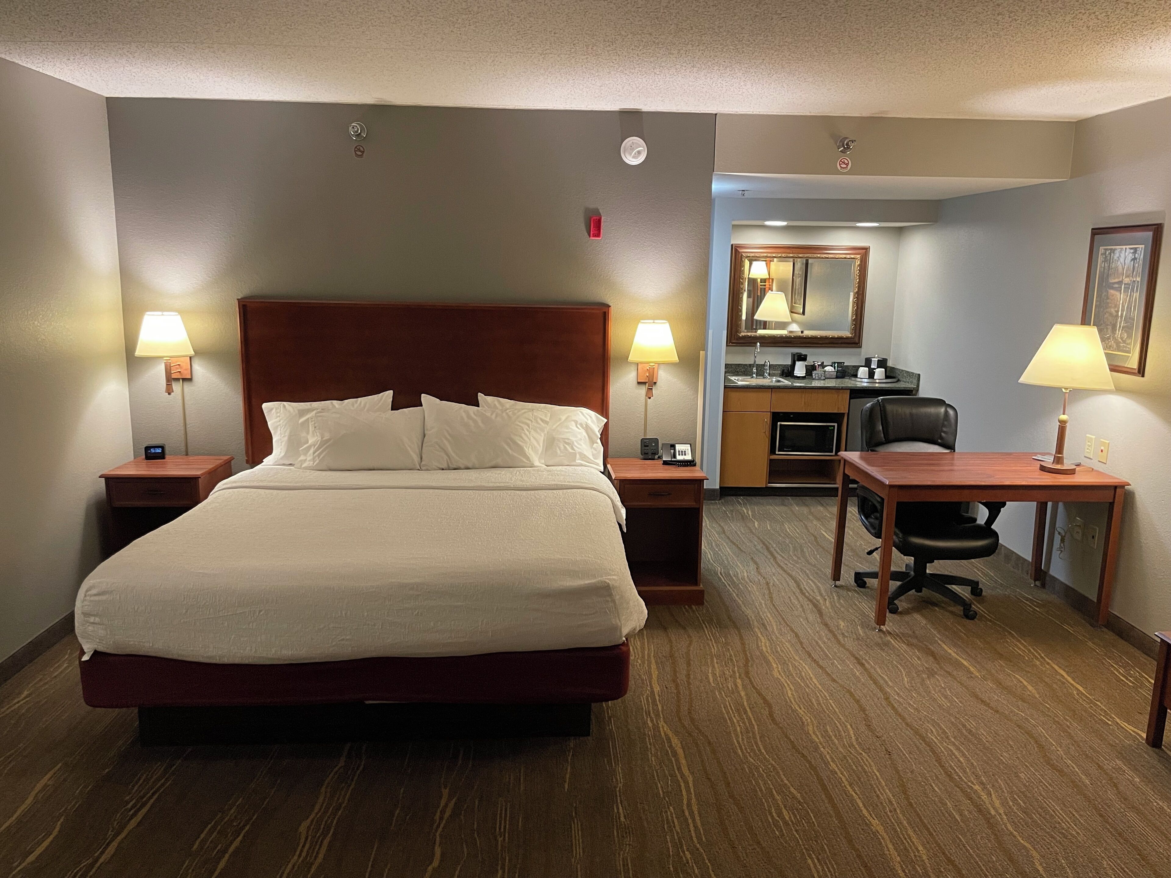 A hotel room with a large bed, two bedside tables with lamps, a desk with a chair, a kitchenette, and a mirror.
