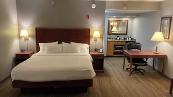 A hotel room with a large bed, two bedside tables with lamps, a desk with a chair, a kitchenette, and a mirror.