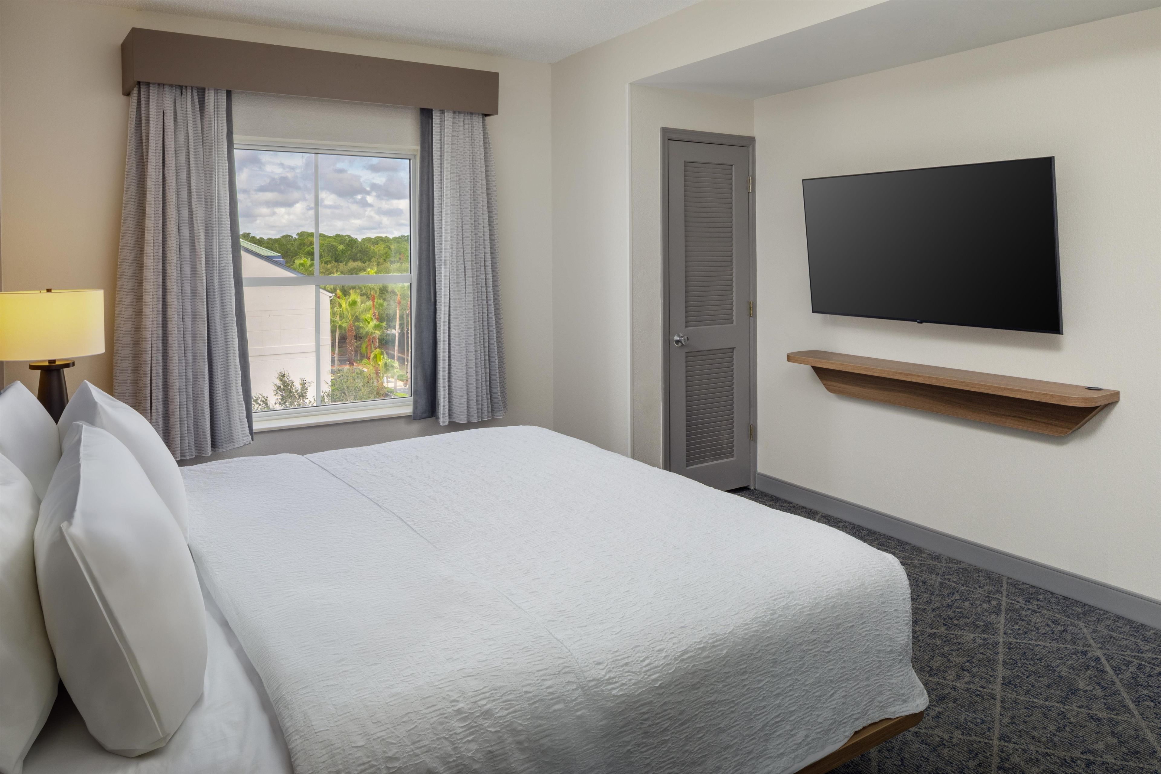 A hotel room with a bed, a TV mounted on the wall, a window with curtains, and a lamp.