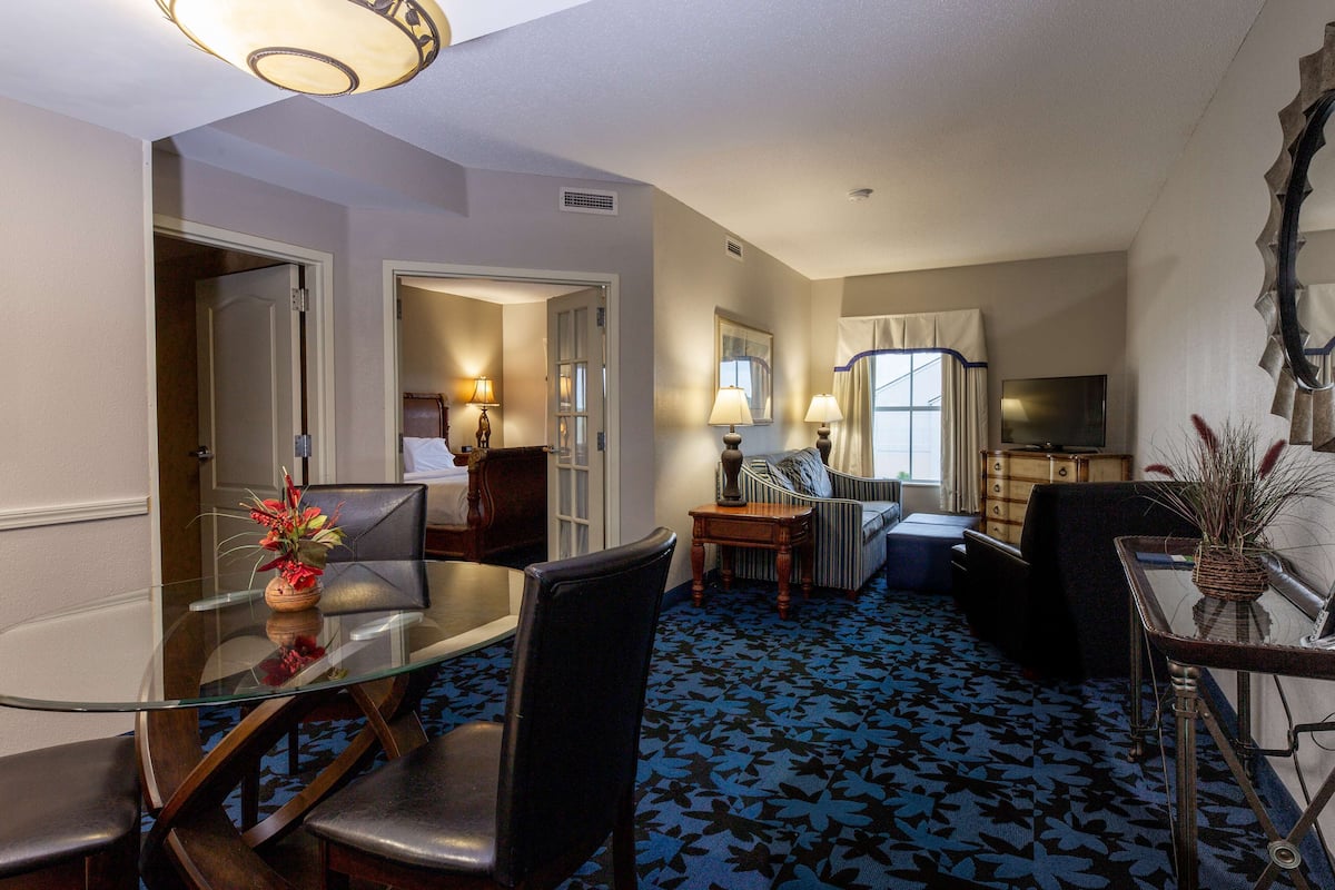A hotel room with a glass dining table, two chairs, a sofa, a TV, and a patterned carpet.