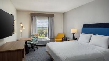 A hotel room with a bed, a desk, a chair, a TV, a window with curtains, and a lamp.