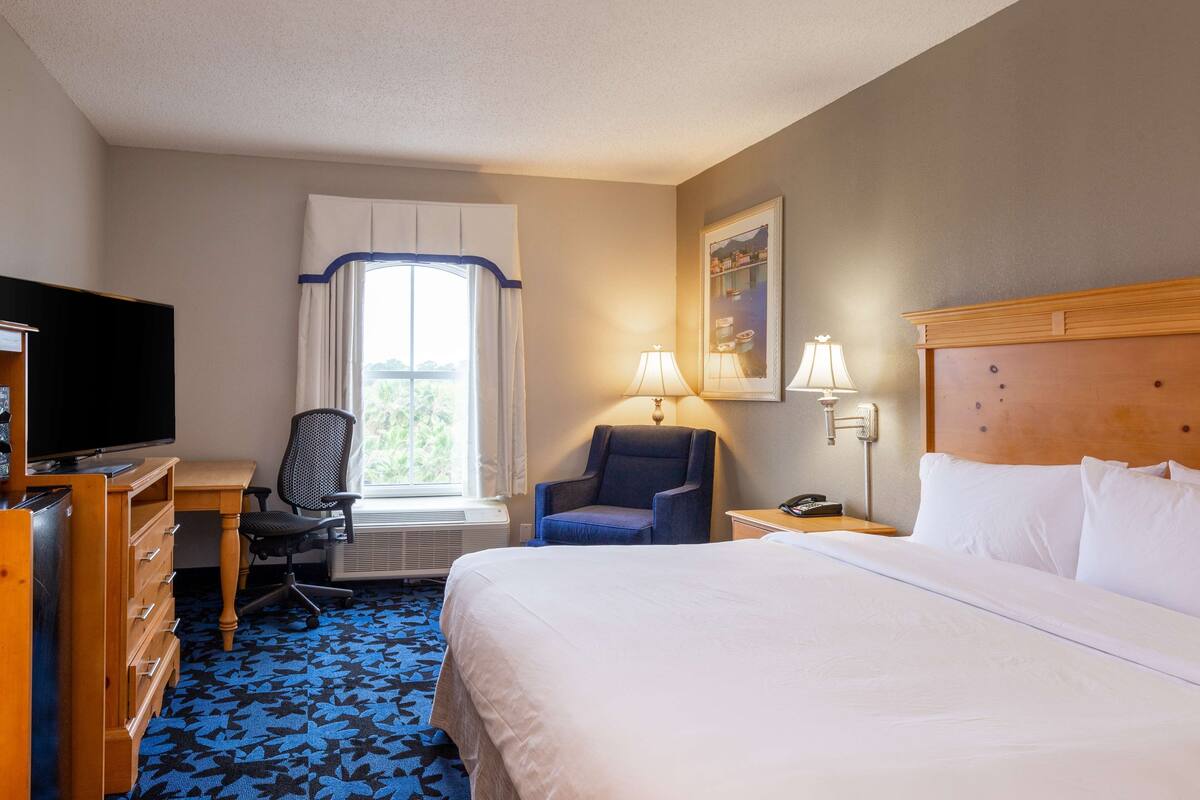 A hotel room with a bed, a desk with a television, a chair, a window with curtains, and a painting on the wall.