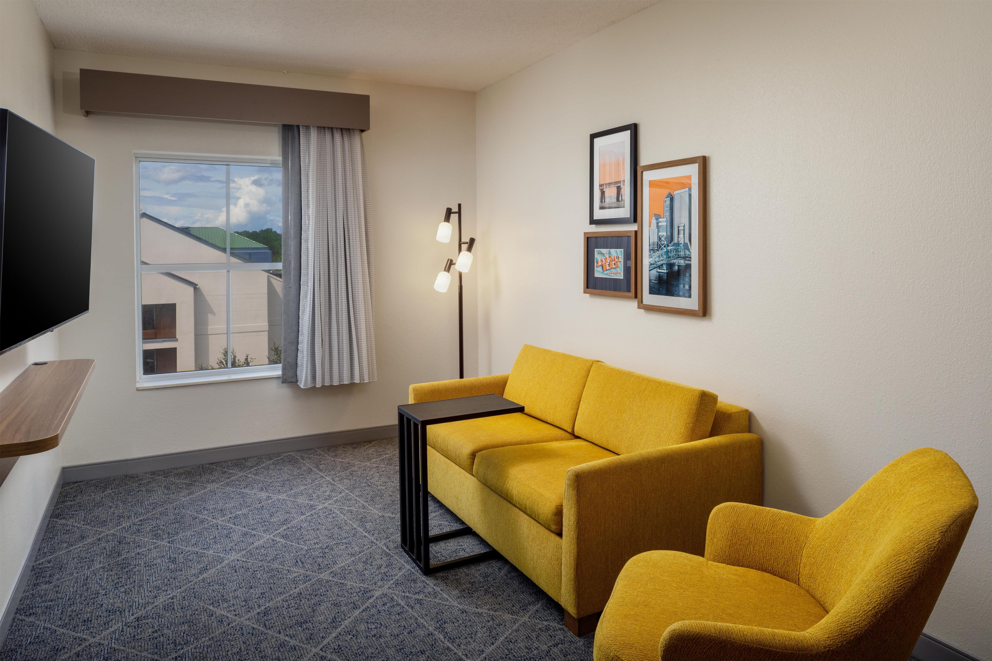 Quarto de hotel com sofá, poltrona, TV de tela plana, janela com cortinas e quadros na parede.