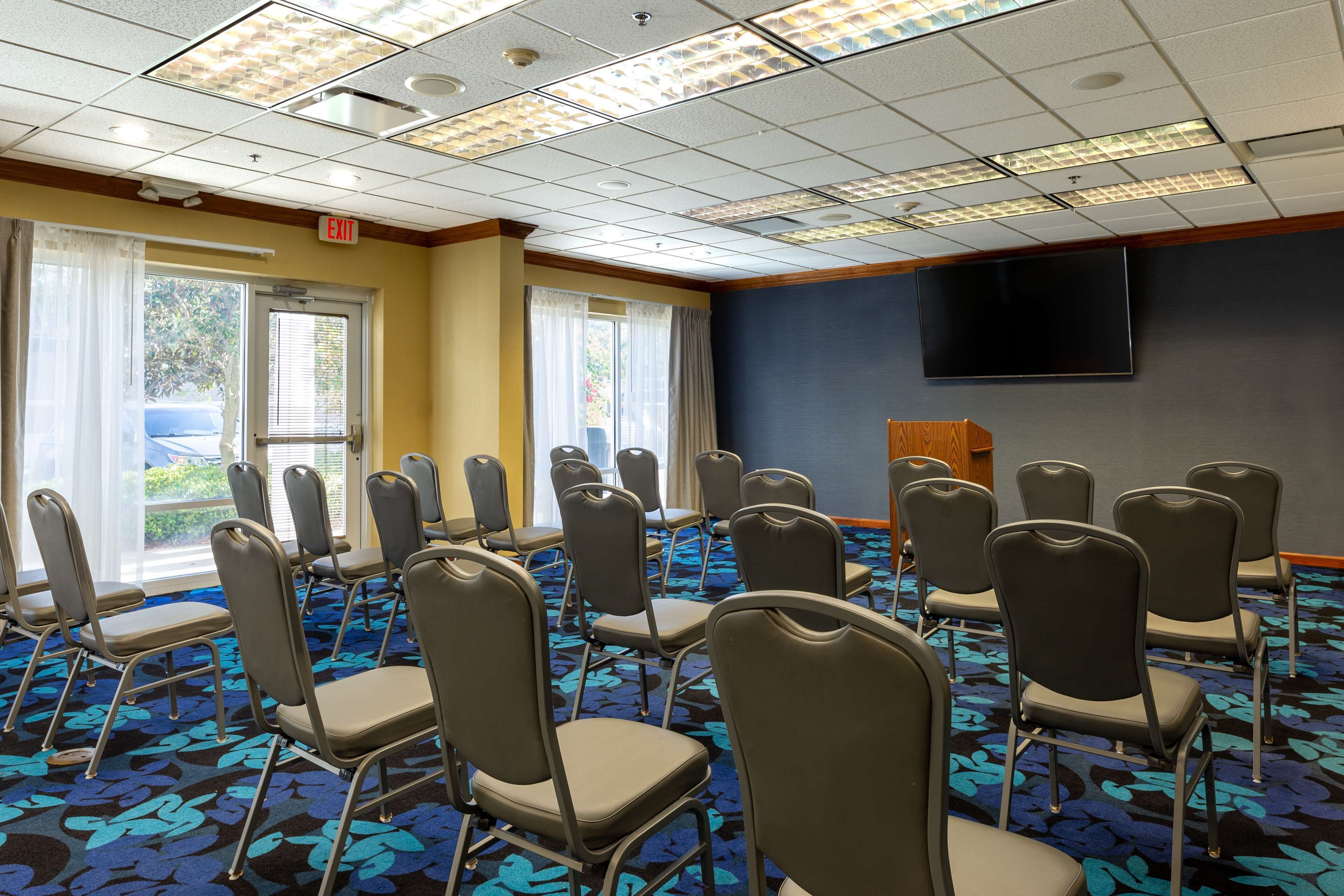 A conference room with rows of chairs, a large flat-screen TV on the wall, and a carpeted floor.