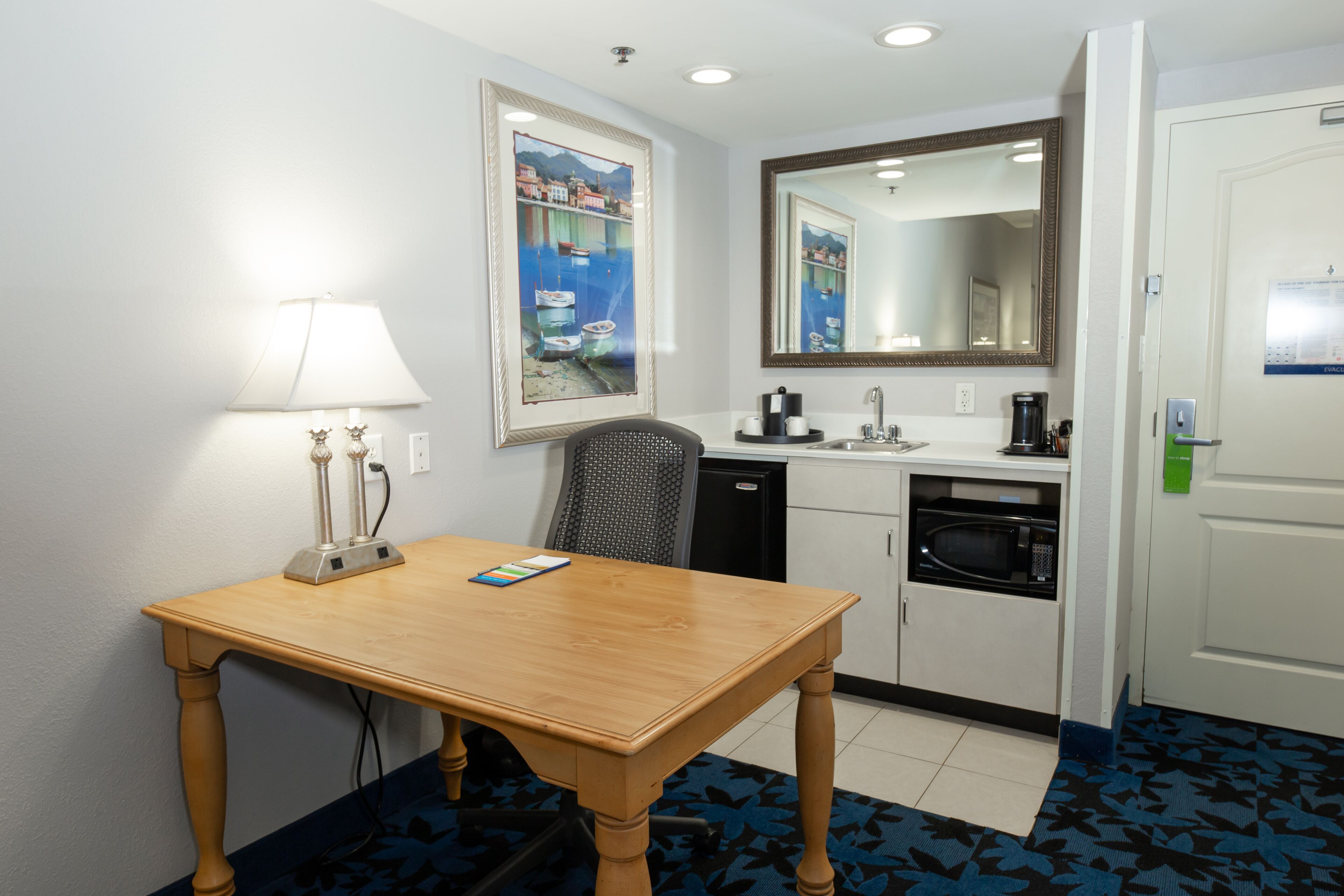 A hotel room with a wooden table, a lamp, a painting, a microwave, and a sink.